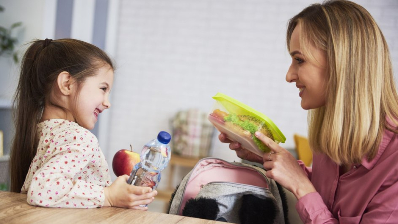 Мама собирает в школу рюкзак дочери и кладет в него ланч-бокс