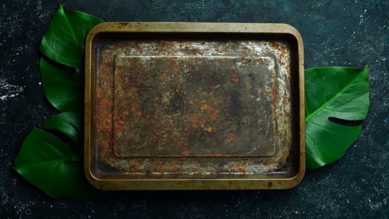 Старый грязный противень лежит на черной столешнице и крупных зеленых листьях